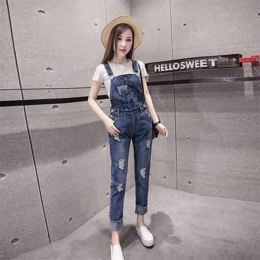 a woman in a hat and overalls stands in front of a wall.