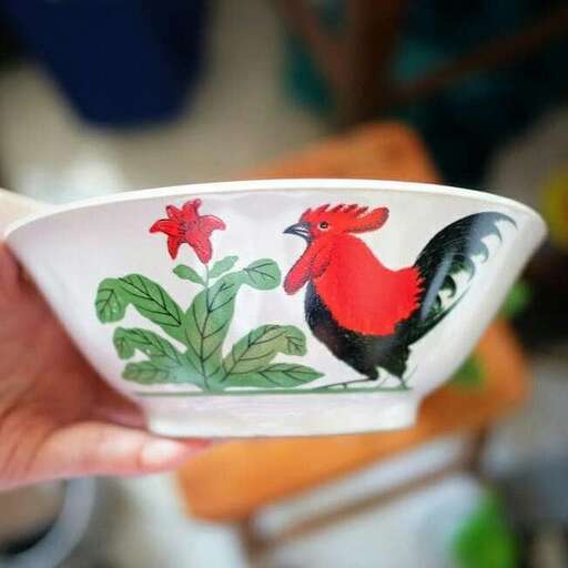 a hand holding a bowl with a rooster on it.